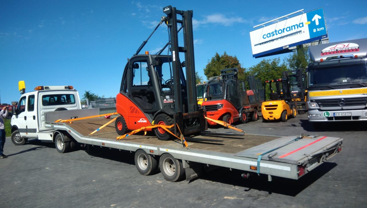 Pomarańczowy wózek widłowy Linde zabezpieczony pasami na platformie lawety, w tle inne wózki widłowe i reklama Castorama.