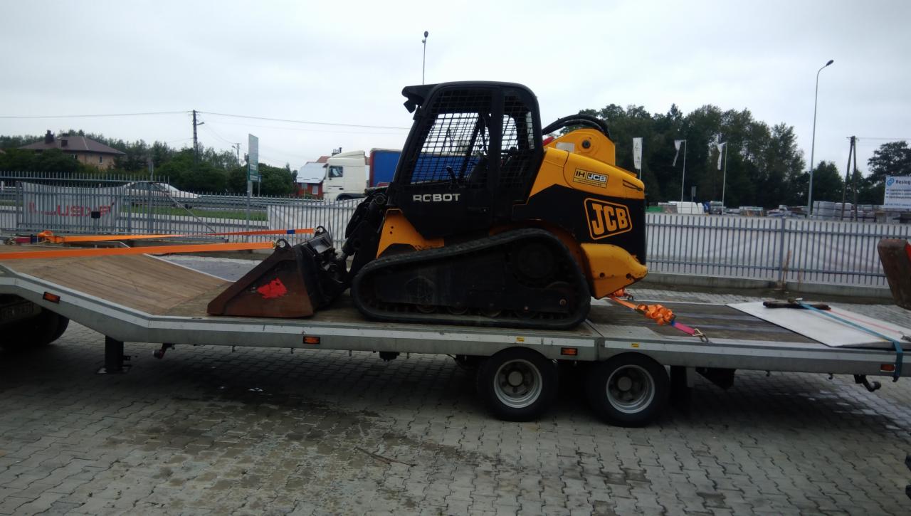 Żółto-czarna miniładowarka gąsienicowa marki JCB typu RC BOT, zabezpieczona pomarańczowymi pasami transportowymi, przewożona na niskopodwoziowej przyczepie lawety, w tle widoczny budynek...