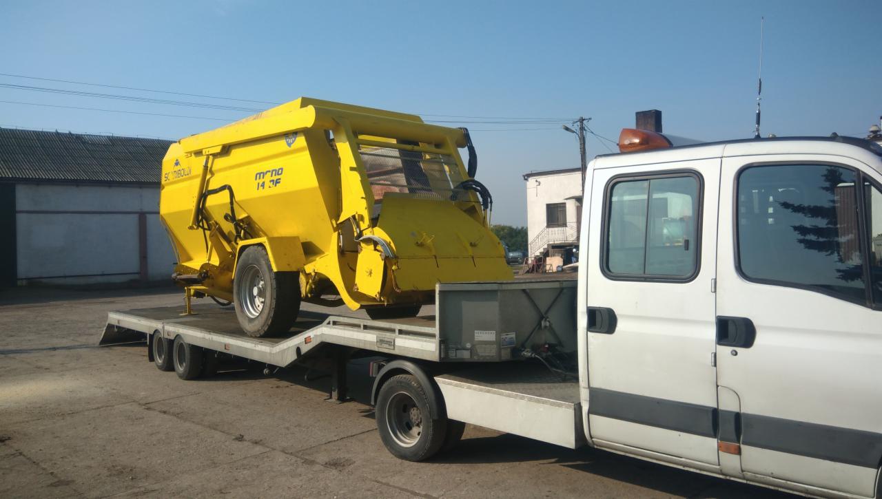 Żółty rozsiewacz nawozów marki Scandibolda model Mono 14 DF przewożony na niskopodwoziowej lawecie ciągniętej przez białego busa dostawczego na tle budynków.