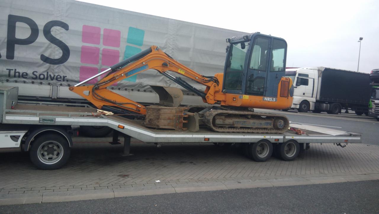 Pomarańczowa mini koparka NR5.5, przewożona na niskopodwoziowej przyczepie, z widocznym logo firmy transportowej na naczepie ciężarówki w tle.
