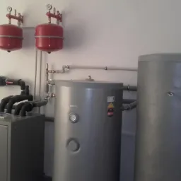 Kompletna instalacja pompy ciepła w kotłowni: szara jednostka wewnętrzna, dwa czerwone zbiorniki wyrównawcze, szare zasobniki ciepłej wody oraz rozdzielacz z czarnymi rurami.