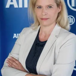 Portret kobiety w beżowej marynarce z założonymi rękami na tle niebieskiej ściany z logo Allianz.