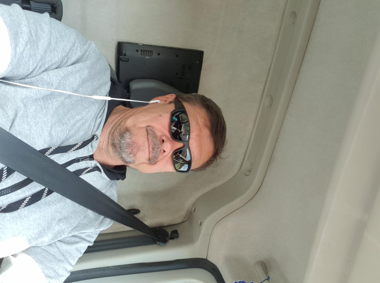 Mężczyzna w okularach przeciwsłonecznych, w samochodzie, z laptopem na siedzeniu obok, podczas podróży.