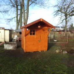 Nowa, pomarańczowa altanka ogrodowa z drewnianymi drzwiami i małym okienkiem, zawieszona doniczka pod okapem, na trawniku w ogrodzie z innymi budynkami w tle.