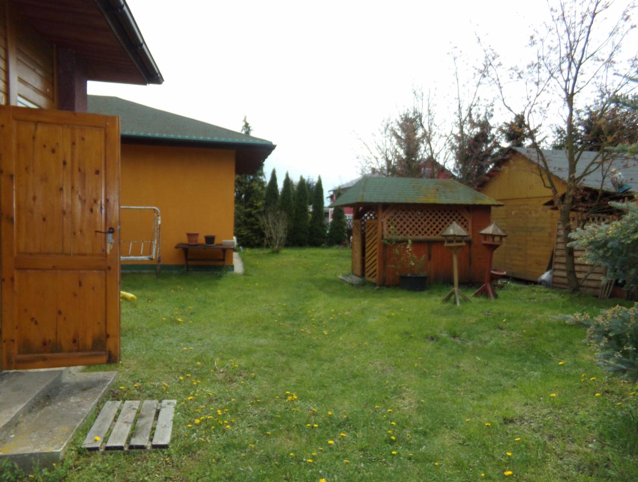 Widok na zadbany ogród z drewnianą altaną z kratownicą, karmnikami dla ptaków, trawnikiem z żółtymi kwiatami i budynkami w tle.