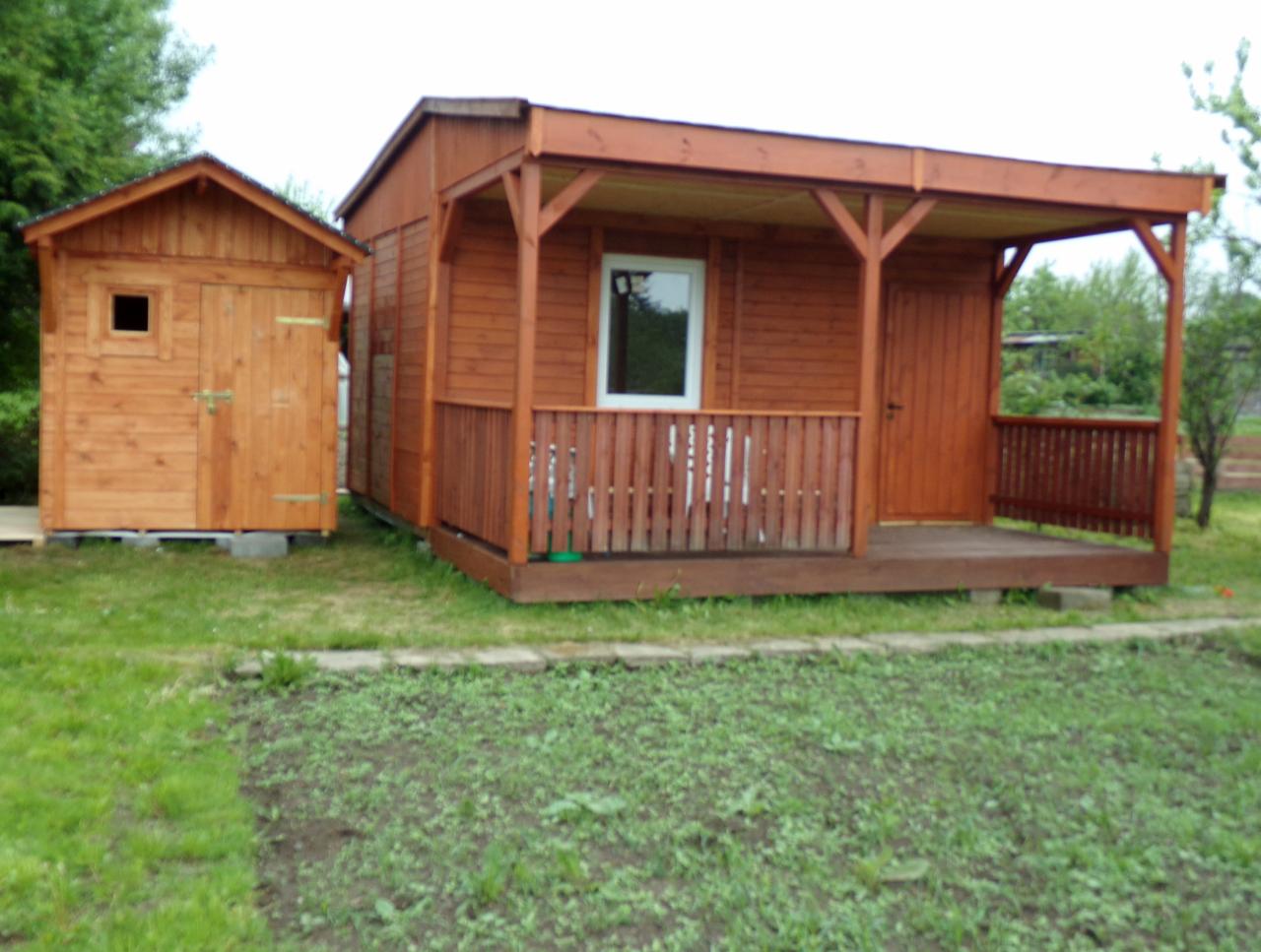 Drewniana altana z zadaszonym tarasem i mała drewniana szopa na trawiastym terenie. Altana ma brązowe deski, białe okno i brązową balustradę. Szopa ma małe okienko i jest umieszczona na betonowych...