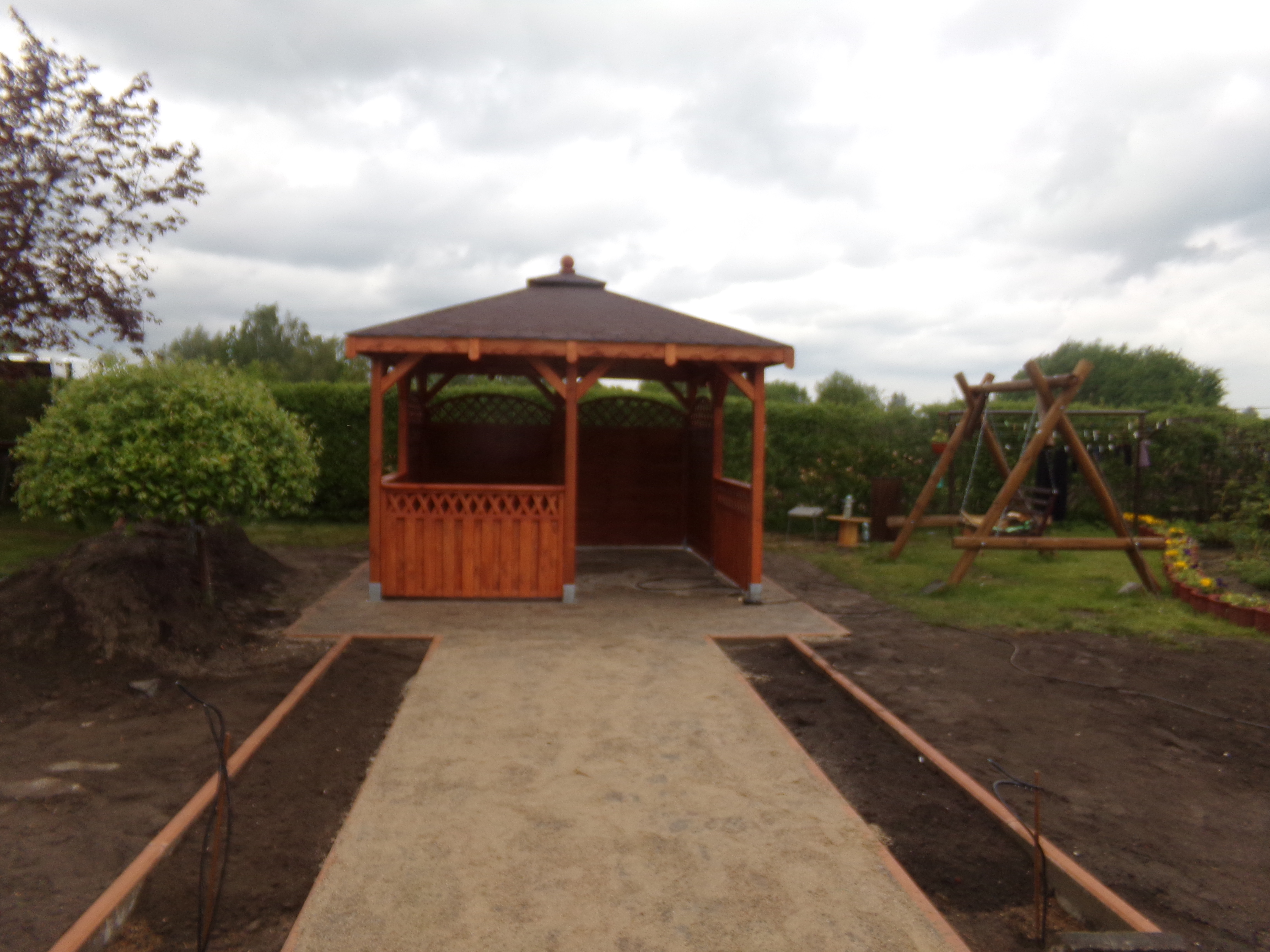 Nowo postawiona drewniana altana z brązowym dachem, widoczna z przodu, z przygotowaną ścieżką z piasku i obramowaniem rabat. W tle huśtawka i drzewa.