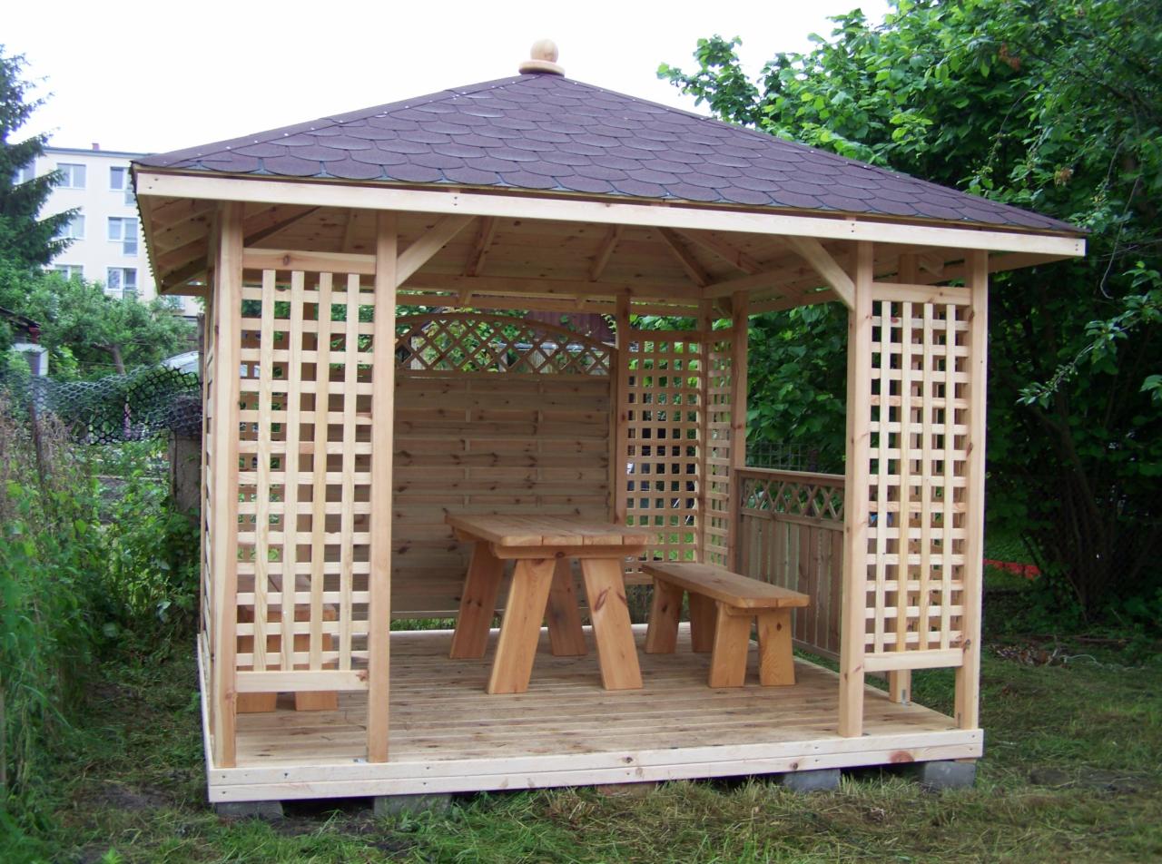 Nowa, surowa altana z jasnego drewna z ażurowymi ścianami, kryta ciemną dachówką, z drewnianym stołem i ławką w środku, ustawiona na trawniku.