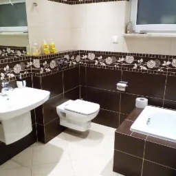 Wykończona łazienka z brązowymi i beżowymi płytkami, dekoracyjnym pasem z motywem róż, wiszącą toaletą i podwieszaną umywalką.