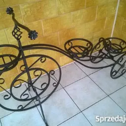 Ozdobny, metalowy kwietnik w kształcie roweru, z trzema miejscami na doniczki. Dekoracyjny element z motywem kwiatowym. Ustawiony na jasnych płytkach, tło stanowi żółta ściana.