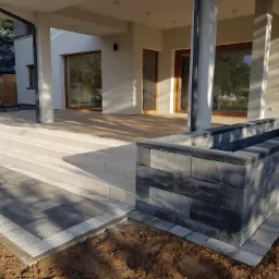 Wykończona ścieżka z kostki brukowej w odcieniach szarości z obramowaniem z jasnej kostki, prowadząca do schodów i tarasu nowoczesnego domu z jasną elewacją i dużymi oknami.