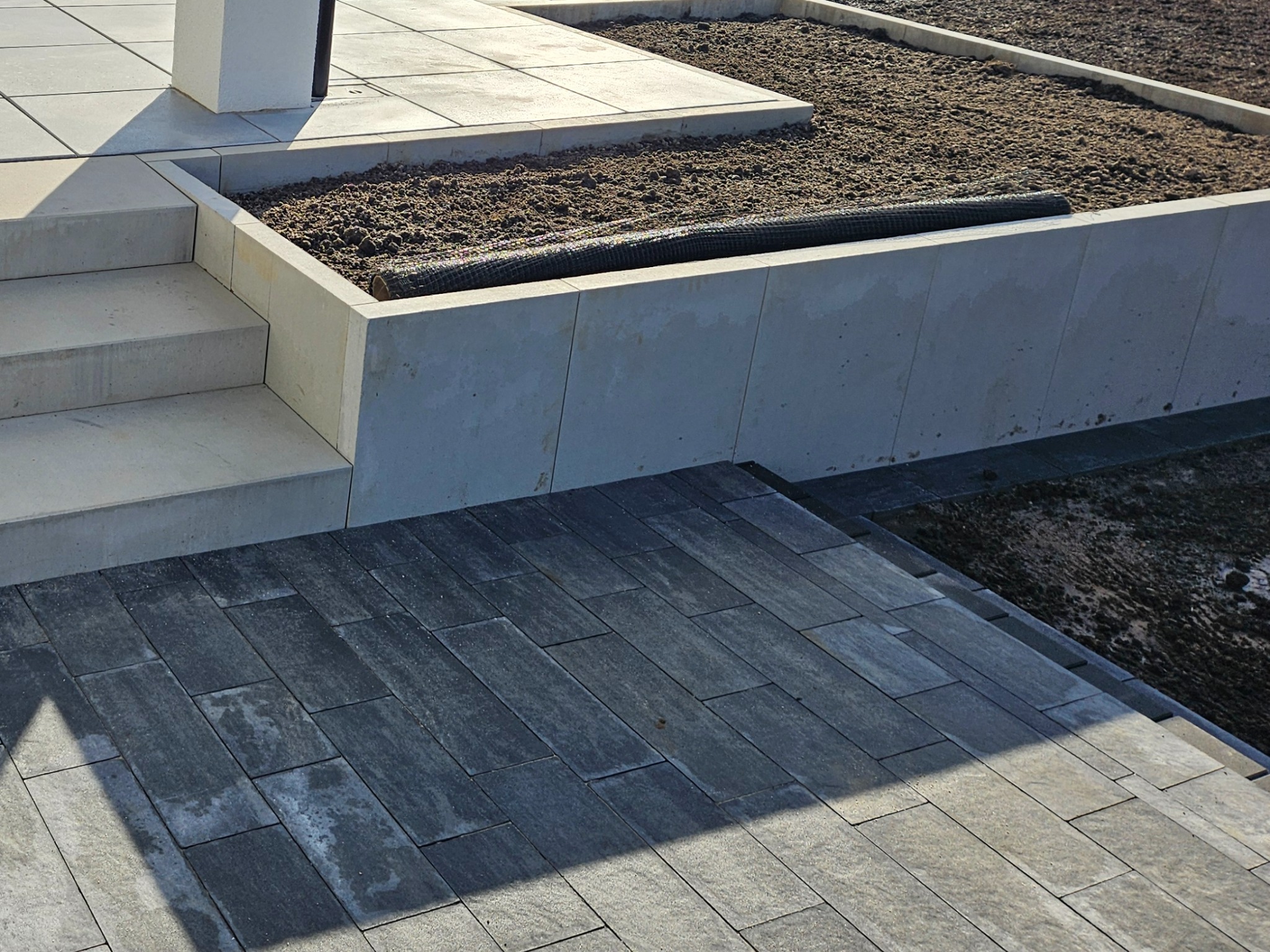 Nowoczesny taras z szarej kostki brukowej ułożonej w prostokątnym wzorze, z niskimi schodami i podwyższonym rabatem z ziemią przygotowaną pod nasadzenia.