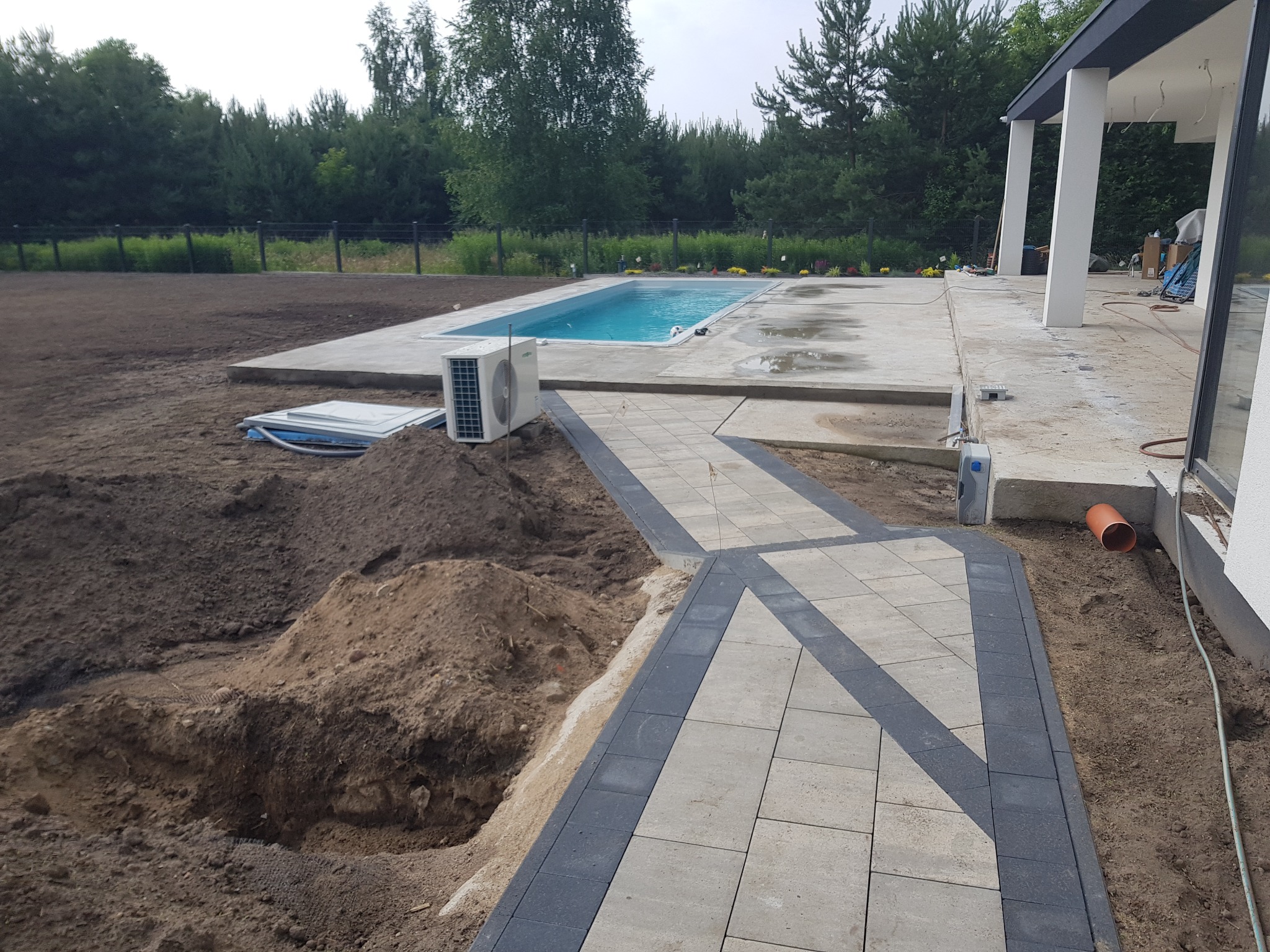 Ścieżka z kostki brukowej w odcieniach szarości prowadzi do basenu w ogrodzie. Widoczny wykop ziemi i klimatyzator. W tle drzewa i nowoczesny dom.