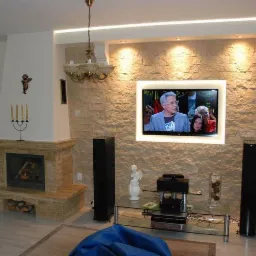 Salon z kominkiem obłożonym kamieniem dekoracyjnym, ściana z telewizorem podświetlona ledami, szklany stolik RTV i kolumny głośnikowe