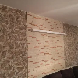 Ściana w salonie z dekoracyjnym tynkiem strukturalnym i panelami imitującymi cegłę, z białą półką zamontowaną na panelach.