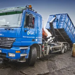 Niebieska ciężarówka Mercedes Actros z kontenerem hakowym podniesionym do góry, widoczne butle z gazem i błotniste podłoże.
