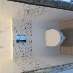 Wykończona toaleta z podwieszaną muszlą, białe i wzorzyste płytki na ścianach, chromowany przycisk spłukiwania. Widok z góry na wnętrze w trakcie wykańczania.