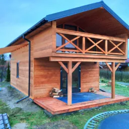Dwukondygnacyjny domek z drewna z balkonem i małym tarasem, stos drewna przy wejściu, widok na trampolinę w ogrodzie.
