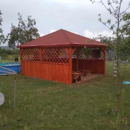 Nowa, drewniana altana z czerwoną dachówką w ogrodzie, z widocznym basenem w tle, wykonana z kratownic i desek.