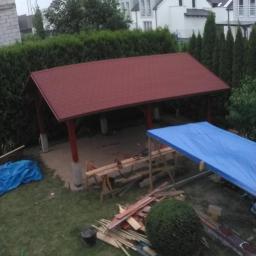 Zygmunt Zumowicz - Wznoszona konstrukcja z czerwonym dachem w ogrodzie, widoczne drewniane elementy konstrukcyjne, narzędzia i plandeki zabezpieczające, w tle domy jednorodzinne i zielona roślinność.