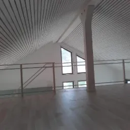 Poddasze z drewnianą podłogą, skośnym sufitem wykończonym panelami i drewnianą balustradą, widok na okna.