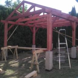Zygmunt Zumowicz - Konstrukcja drewnianej altany w trakcie budowy, widoczne betonowe podstawy, rusztowanie i drabina, w tle zieleń.