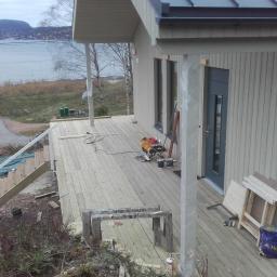 Zygmunt Zumowicz - Budowa drewnianego tarasu przy domu z widokiem na jezioro, widoczne narzędzia i materiały budowlane, częściowo ukończone schody.