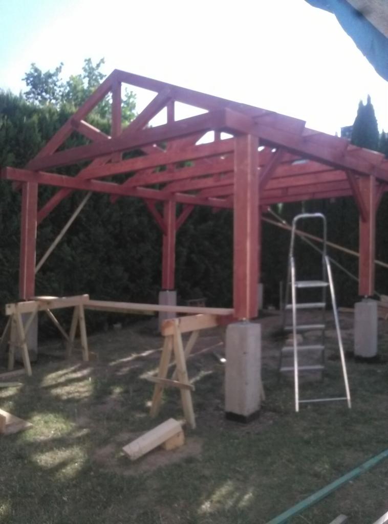 Konstrukcja drewnianej altany w trakcie budowy, widoczne betonowe podstawy, rusztowanie i drabina, w tle zieleń.