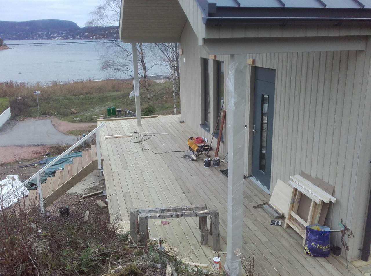 Budowa drewnianego tarasu przy domu z widokiem na jezioro, widoczne narzędzia i materiały budowlane, częściowo ukończone schody.