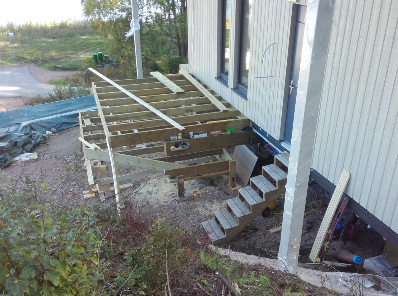 Konstrukcja drewniana pod przyszły taras przy budynku z drewnianą elewacją, widoczne schody wejściowe, teren wokół w trakcie prac ziemnych.