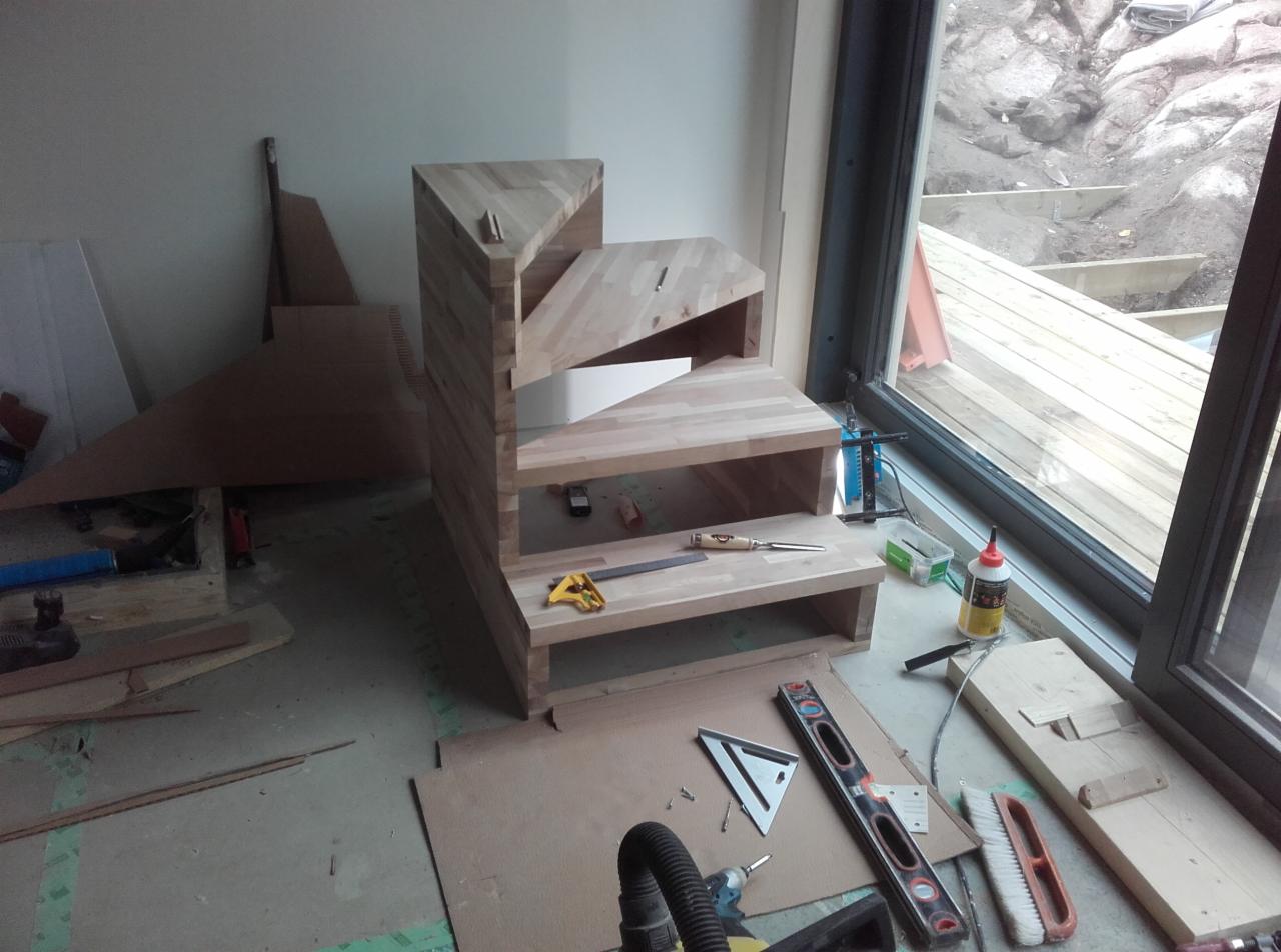 Niedokończona, drewniana konstrukcja schodów z widocznymi narzędziami stolarskimi w pomieszczeniu z dużym oknem, przez które widać fragment tarasu.