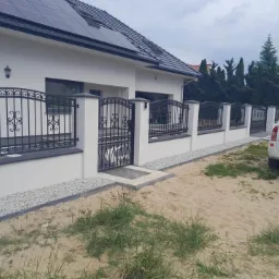 Czarne, dekoracyjne przęsła i brama z elementami kutymi, zamontowane na niskim, białym murku z szarymi słupkami w Zielonej Górze. Widoczny fragment domu z panelami słonecznymi i podjazd z piaskiem...