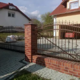 Dwie przesuwne bramy wjazdowe z kutego metalu, osadzone na ceglanych słupkach, prowadzące do posesji z dwoma domami, widoczny samochód w garażu.