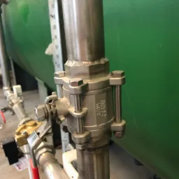 Metalowa armatura z zaworem kulowym na tle zielonego zbiornika, widoczne spawane łączenia rur.