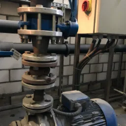 Silnik pompy z rurociągami połączonymi kołnierzami w kotłowni, widoczna armatura, przewody elektryczne i tablica sterownicza na tle ściany z białej cegły.