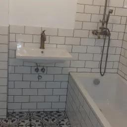 Biała łazienka z wanną i umywalką, wykończona białymi płytkami z czarną fugą oraz dekoracyjną podłogą w stylu retro, bateria umywalkowa i prysznicowa w kolorze starego złota.