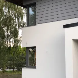 Nowoczesny dom z białymi ścianami i szarą elewacją z desek, widoczne okna z ciemnymi ramami i fragment dachu, otoczony zielenią.