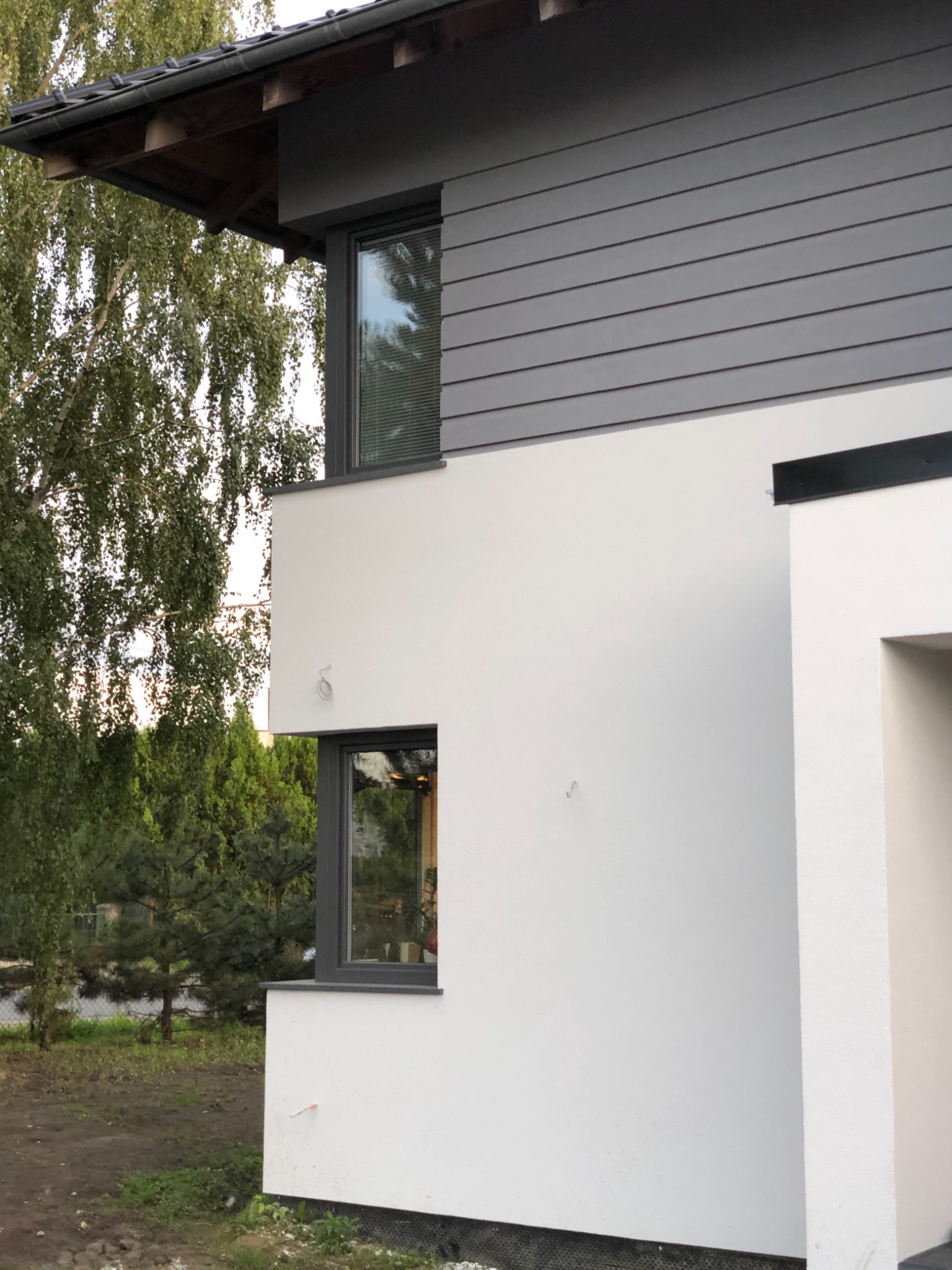 Nowoczesny dom z białymi ścianami i szarą elewacją z desek, widoczne okna z ciemnymi ramami i fragment dachu, otoczony zielenią.