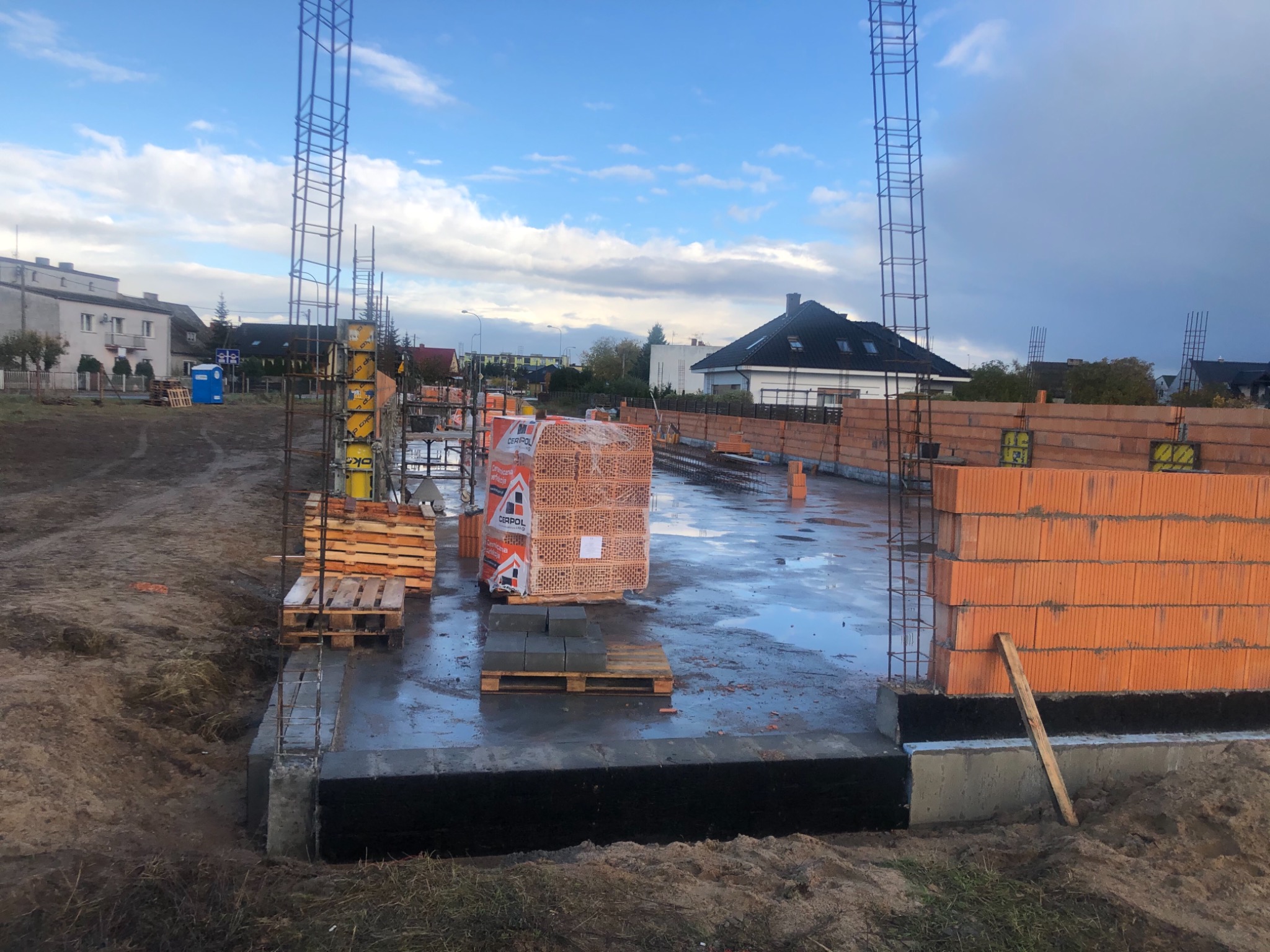 Budowa domu jednorodzinnego: wylana płyta fundamentowa, wzniesione fragmenty ścian z czerwonej cegły ceramicznej, widoczne zbrojenia pionowe, palety z cegłami CERPOL na mokrej powierzchni, w tle...