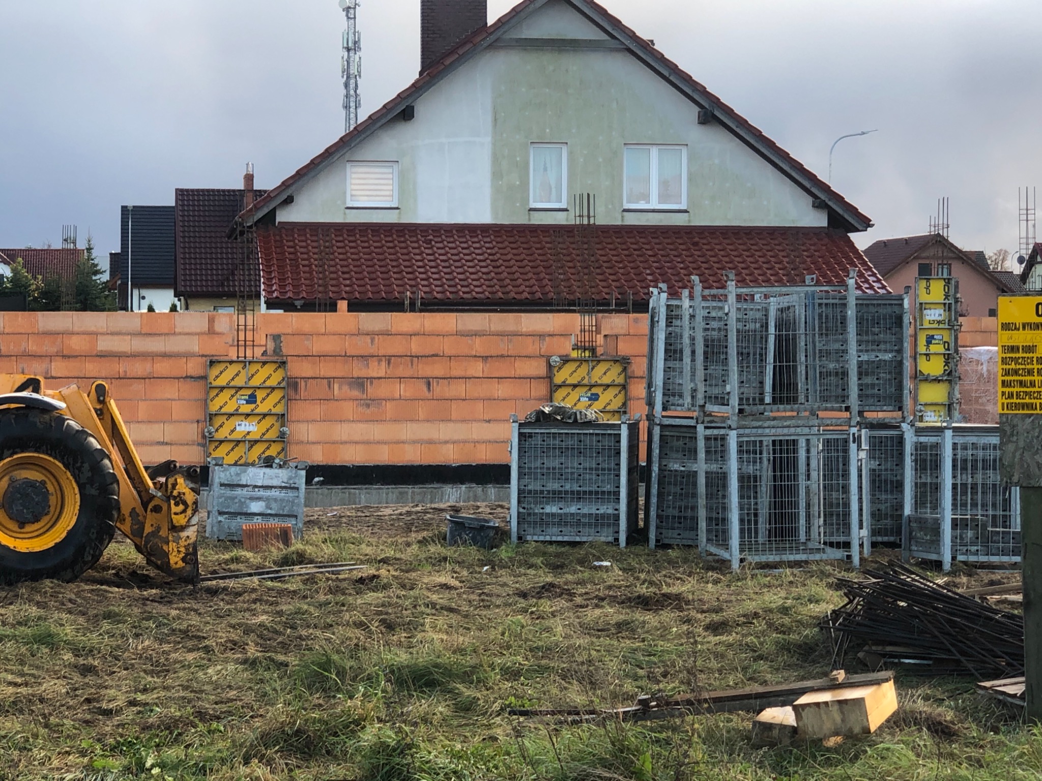 Wznoszenie ściany z czerwonej cegły na budowie domu jednorodzinnego, widoczne szalunki i elementy konstrukcyjne, na pierwszym planie żółta koparka i stosy metalowych klatek.