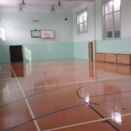 Błyszcząca podłoga w sali gimnastycznej po umyciu, widoczne linie boiska i kosz do koszykówki.