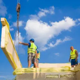 Montaż ściany domu szkieletowego z użyciem dźwigu, pracownicy w kamizelkach odblaskowych i kaskach na tle błękitnego nieba z chmurami.