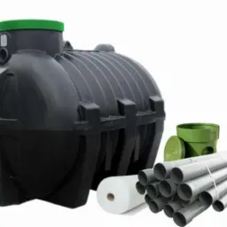 Czarny, plastikowy zbiornik z zieloną pokrywą, rury PCV związane sznurkiem, rolka białej włókniny i zielony element studzienki na białym tle.