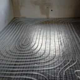 Białe rury ogrzewania podłogowego ułożone w pętle na czarnej siatce montażowej, widok z góry na surowy stan wykończenia pomieszczenia.