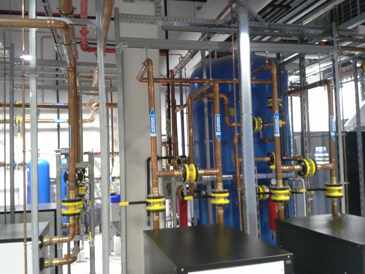Złożona instalacja rur miedzianych z zaworami i zbiornikami w pomieszczeniu technicznym, widoczne metalowe konstrukcje pod sufitem.