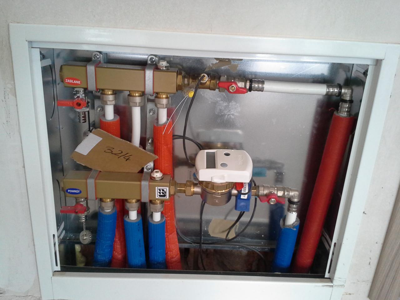 Rozdzielacz instalacji wodnej z licznikiem w zabudowie w Legnicy. Widoczne rury w izolacji termicznej koloru czerwonego i niebieskiego oraz zawory kulowe.