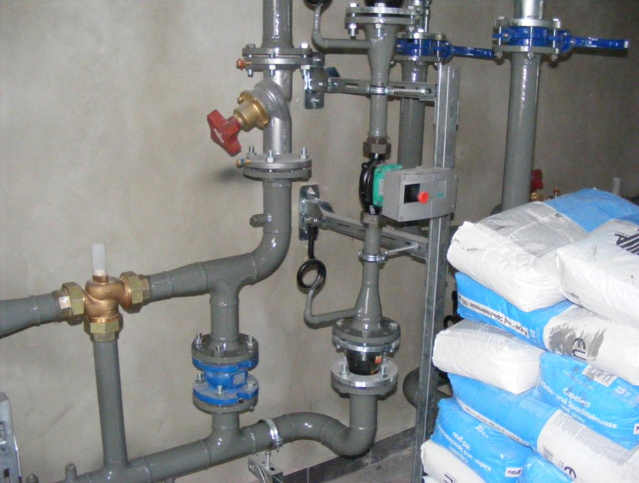 Szare rury instalacji hydraulicznej z zaworami, przyłączeniami kołnierzowymi i pompą obiegową na tle surowej ściany, obok stos worków z zaprawą.