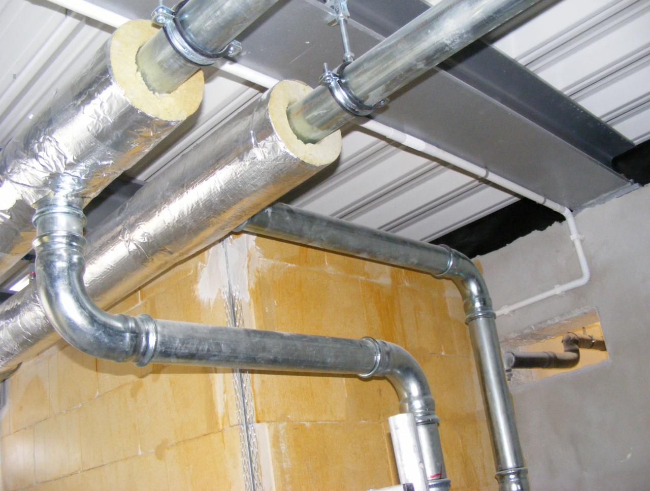 Instalacja rur z izolacją termiczną, mocowanych do sufitu i ściany z pustaków, widoczne złącza i fragment instalacji w otworze w ścianie.