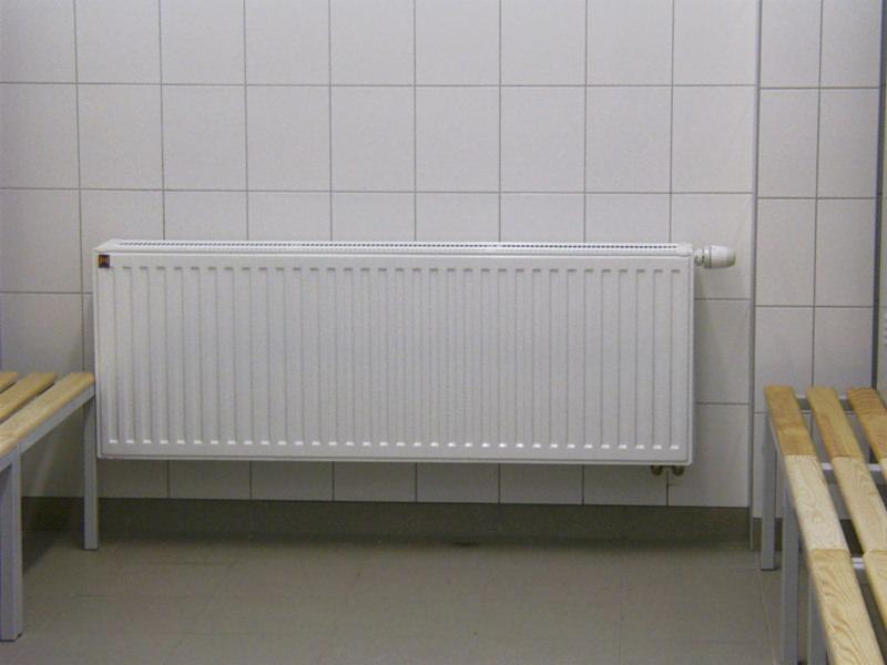 Biały grzejnik panelowy zamontowany na ścianie wyłożonej białymi płytkami w szatni z drewnianymi ławkami.