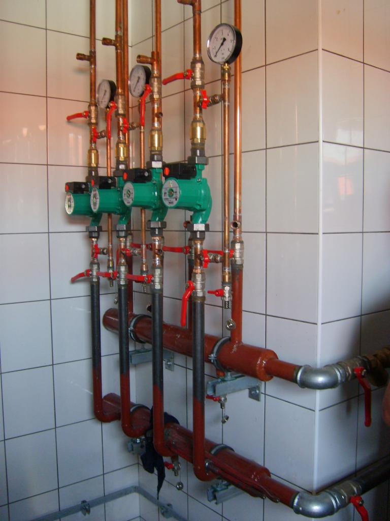 Instalacja centralnego ogrzewania z trzema zielonymi pompami obiegowymi, manometrami i zaworami, zamontowana na tle białych, kwadratowych płytek.